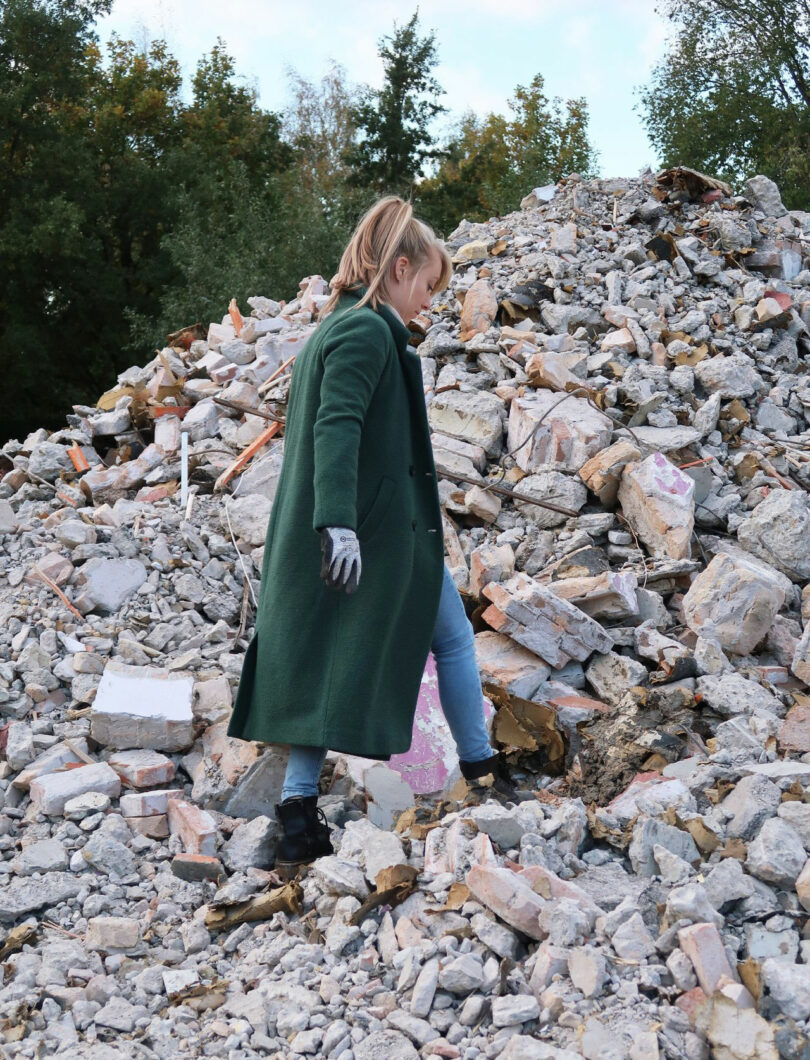 A person in a green coat and gloves walks on a pile of rubble and broken bricks outdoors, with trees visible in the background, while a curious Jule Cats figure peeks out from behind the debris.