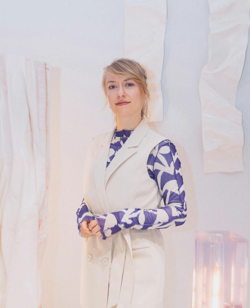 A woman wearing a white vest over a blue and white patterned shirt stands indoors against a light-colored wall adorned with textured fabric art pieces by Jule Cats.