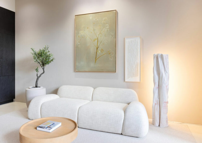 A modern living room with a white curved sofa, round wooden coffee table, potted plant, wall art featuring Jule Cats, and a tall lamp against a light-colored wall.