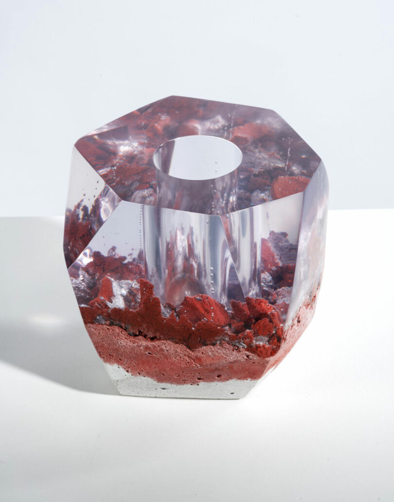 A geometric, transparent resin sculpture by Jule Cats features a central hollow and red, rock-like material embedded at the base, displayed on a white surface.