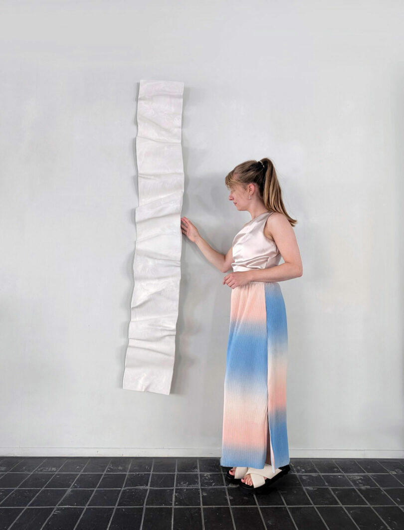 A woman in a satin top and gradient skirt stands indoors, touching a tall, narrow, white sculptural piece by Jule Cats mounted on a light gray wall.