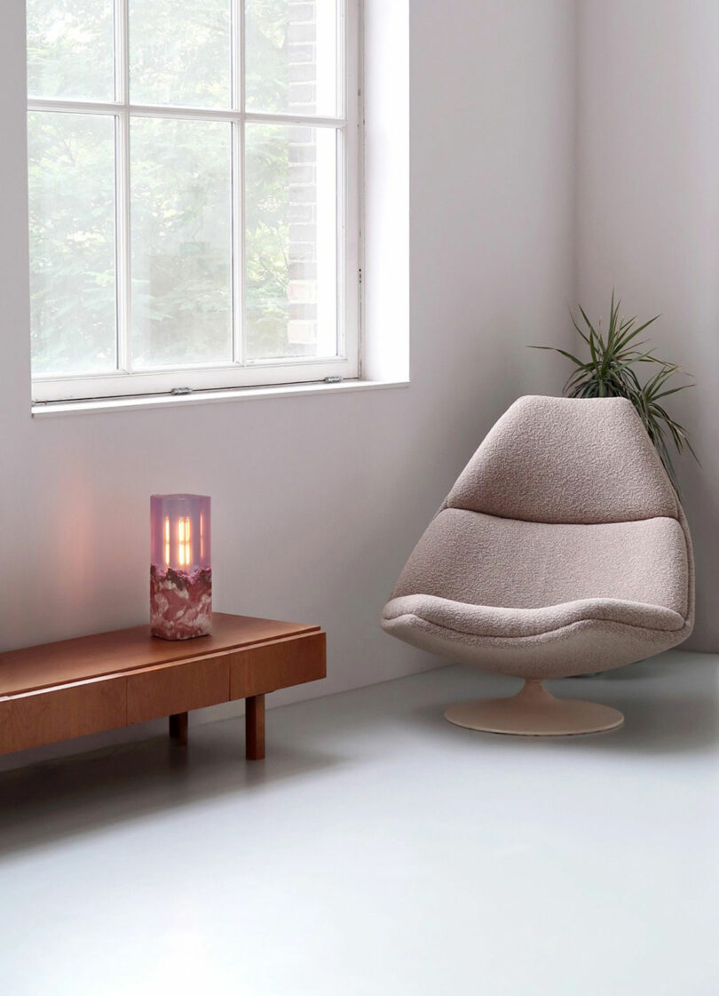 A modern Jule Cats lounge chair sits in a minimalist room next to a low wooden table with a lamp, a potted plant, and a large window letting in natural light.