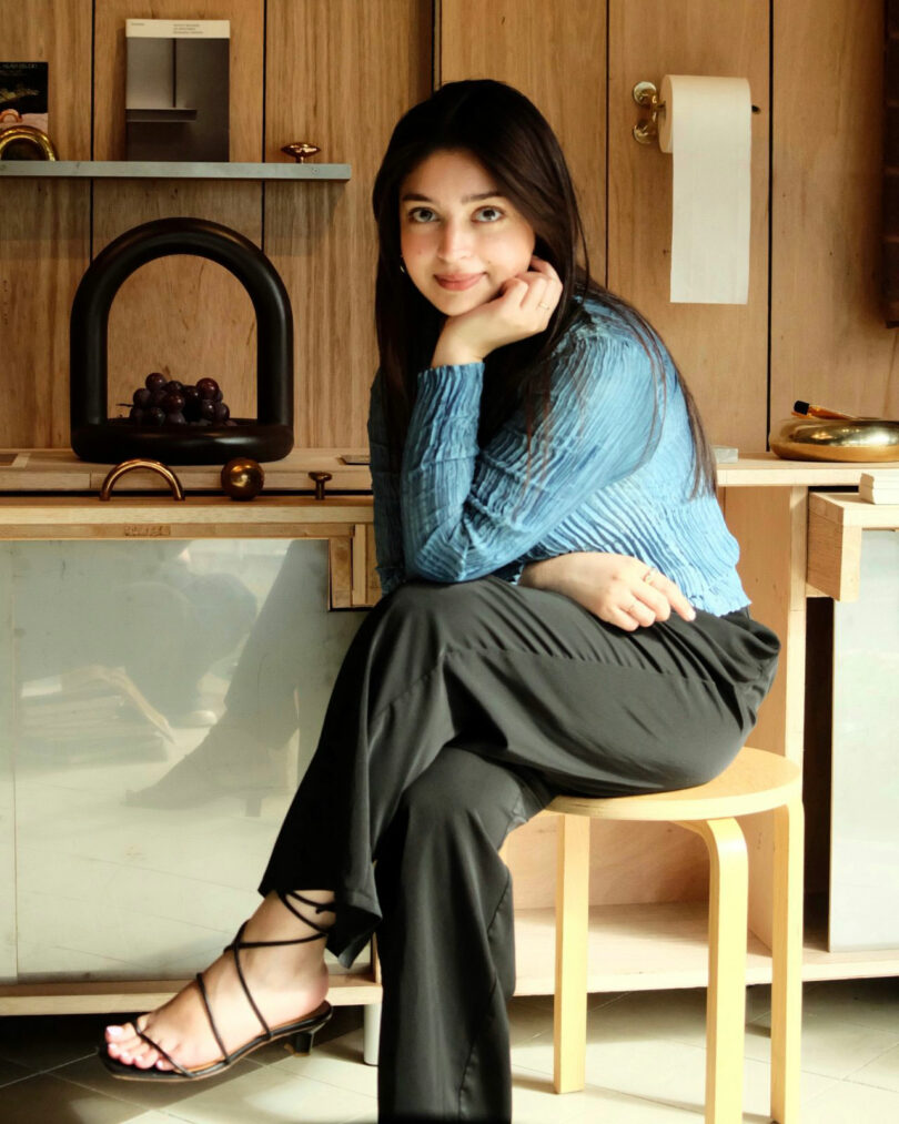 Maha Alavi, with long dark hair, wearing a blue blouse and black pants, sits on a wooden stool in a modern kitchen, resting her chin on her hand and looking at the camera.