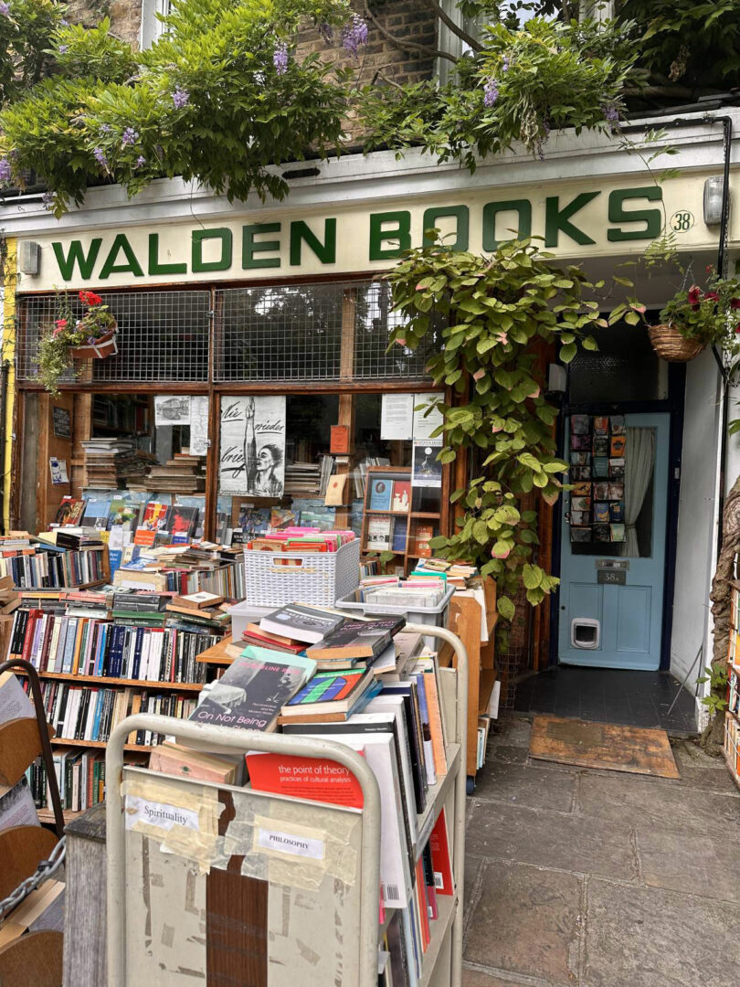 The exterior of Walden Books, designed by Maha Alavi, features bookshelves and carts of books displayed outside, surrounded by greenery and potted plants.