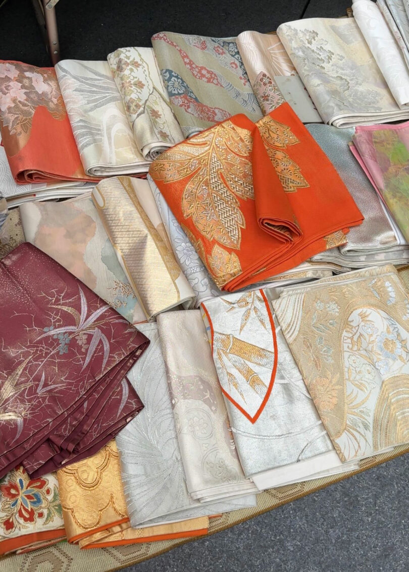 Several folded pieces of ornate, patterned fabric by Maha Alavi in vibrant hues—orange, maroon, gold, and cream—are elegantly displayed on a flat surface.