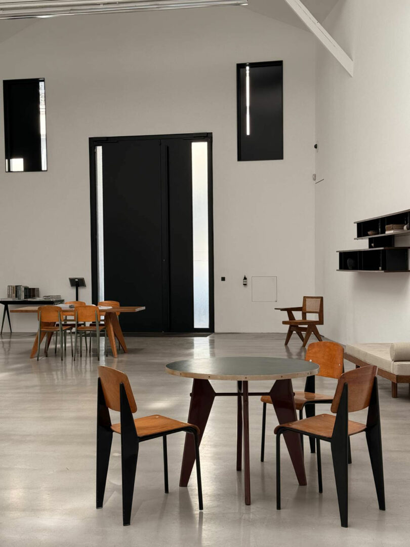 Minimalist interior inspired by Maha Alavi, featuring round and rectangular tables, wooden chairs, a large black door, high windows, and wall-mounted shelves in a spacious, well-lit room.