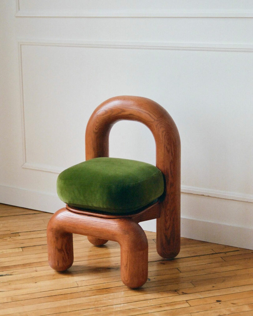 A modern chair with a rounded wooden frame and legs, featuring a curved backrest and a green velvet seat cushion, inspired by Maha Alavi’s design aesthetic, placed on a hardwood floor against a white wall.