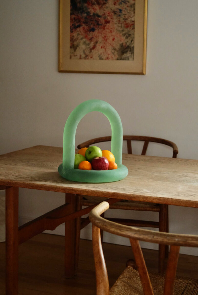 A modern green fruit holder with an arch design sits on a wooden table, containing apples, oranges, and lemons; a framed abstract painting by Maha Alavi hangs on the wall behind.