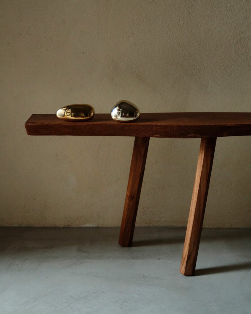 A wooden bench with three angled legs stands against a plain wall, featuring two polished, rounded metal objects on top—a minimalist design inspired by Maha Alavi.