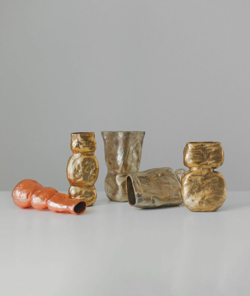 Five abstract, irregularly shaped sculptural vessels by Maha Alavi in metallic gold and bronze tones, and one in orange, are displayed on a plain white surface against a neutral background.