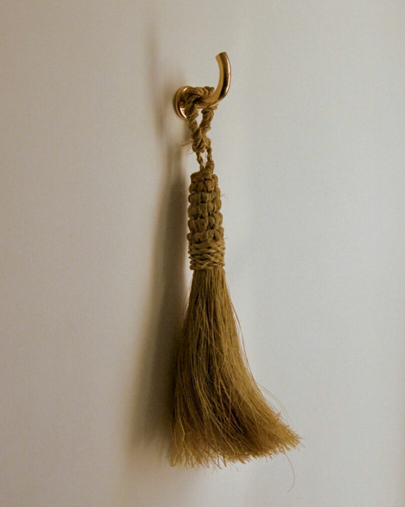 A bundle of coarse brown fibers tied with braided string hangs from a metal hook against a plain wall, evoking the tactile minimalism often seen in Maha Alavi's work.