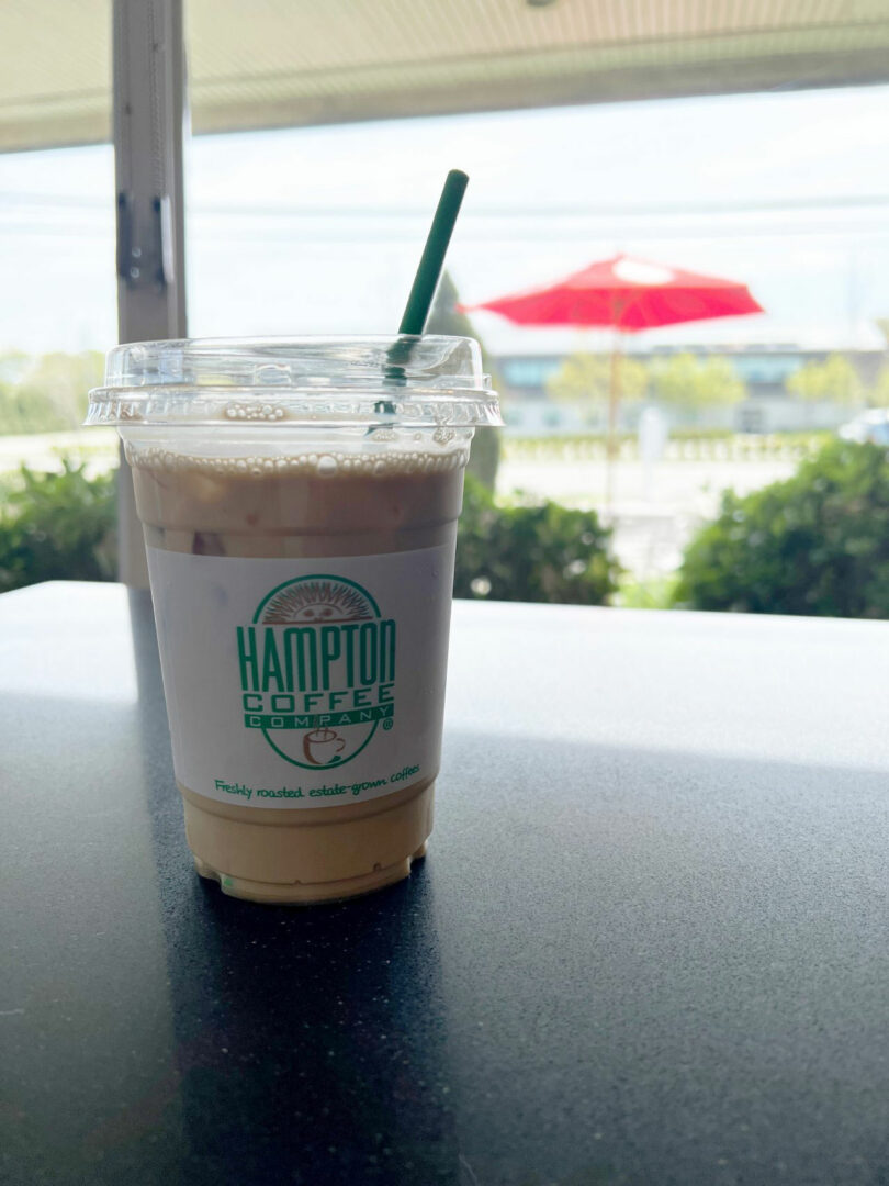 Plastic cup of ice coffee with green straw from Hampton Coffee Company sits on the table; On the open scene with a red umbrella, it was visible in the background, captured Nicki Gitlin.