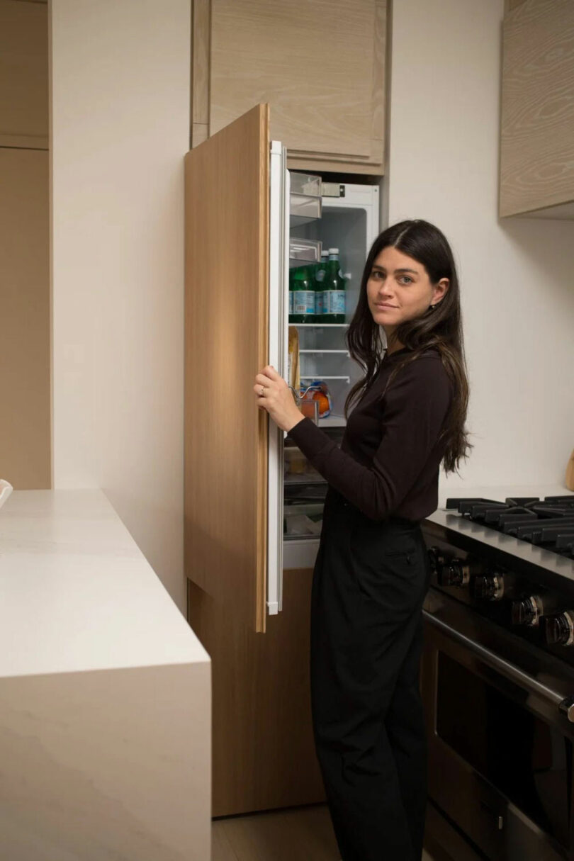 Nicki Gitlin stands in the kitchen, opening the door of the refrigerator and looking at the camera. The refrigerator contains different items, and the furnace is visible next to it.
