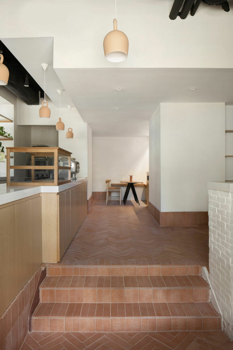 The minimalist interior of the Kafe Nicki Gitlin has floors for Terracotta tiles, lightly wooden cabinet, pendant lights and a small dining room with table and stools in the background.