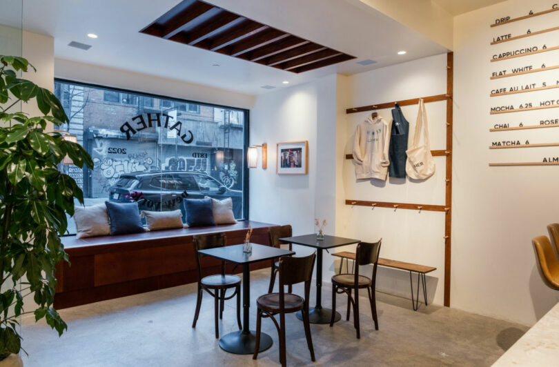 Modern coffee interior Nicki Gitlin with wooden tables and chairs, benches with a large window, hooks with coats with bags, wall art and match plants. Street and parked cars are visible outside.