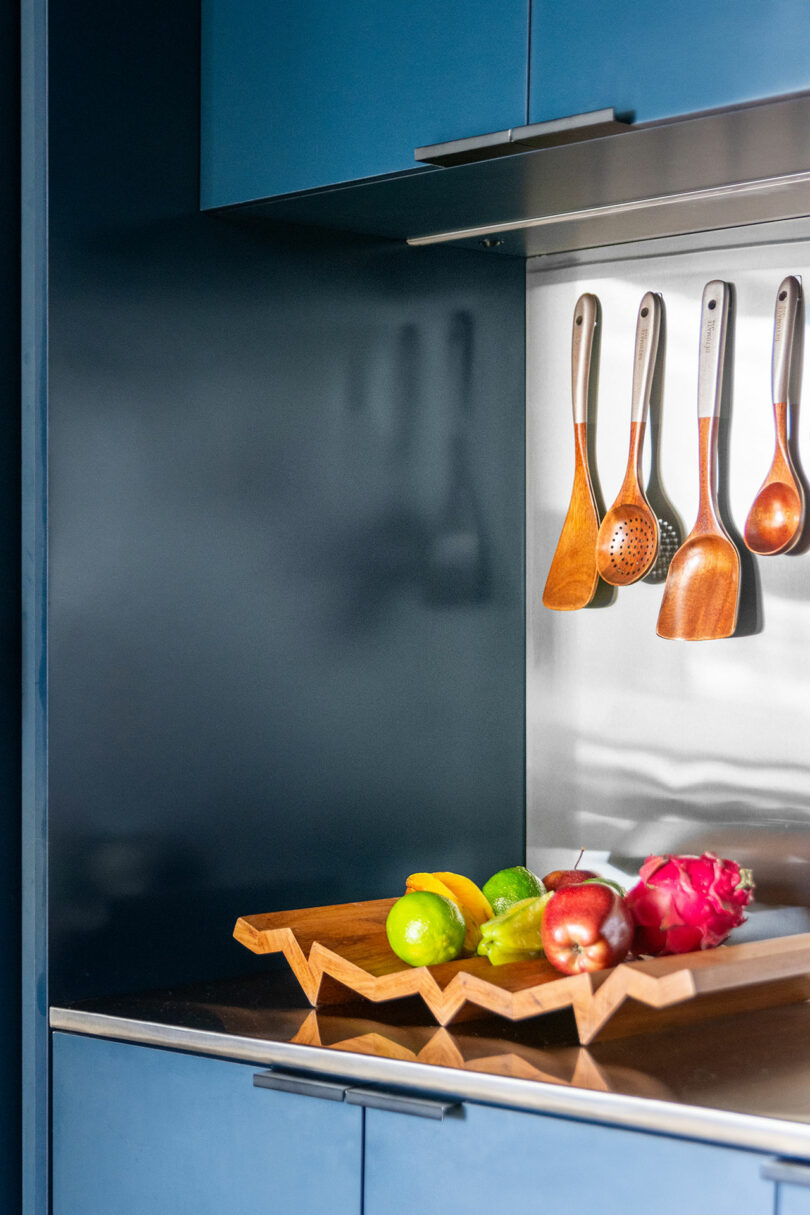 Una cocina moderna con utensilios de madera colgando de una rejilla y una bandeja de madera geométrica que contiene varias frutas en el mostrador.