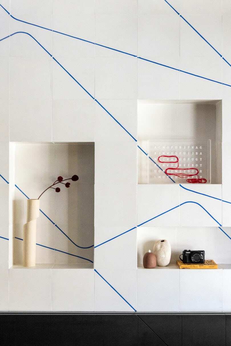Una pared de azulejos blancos con líneas azules presenta estantes empotrados que sostienen un jarrón con tallos rojos, un reloj de palabras, jarrones decorativos, una cámara y libros amarillos.