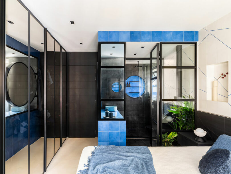 Baño moderno con azulejos negros y azules, particiones de vidrio, espejos redondos, una pequeña zona de fregadero y una planta verde al lado de una cama con una manta azul.