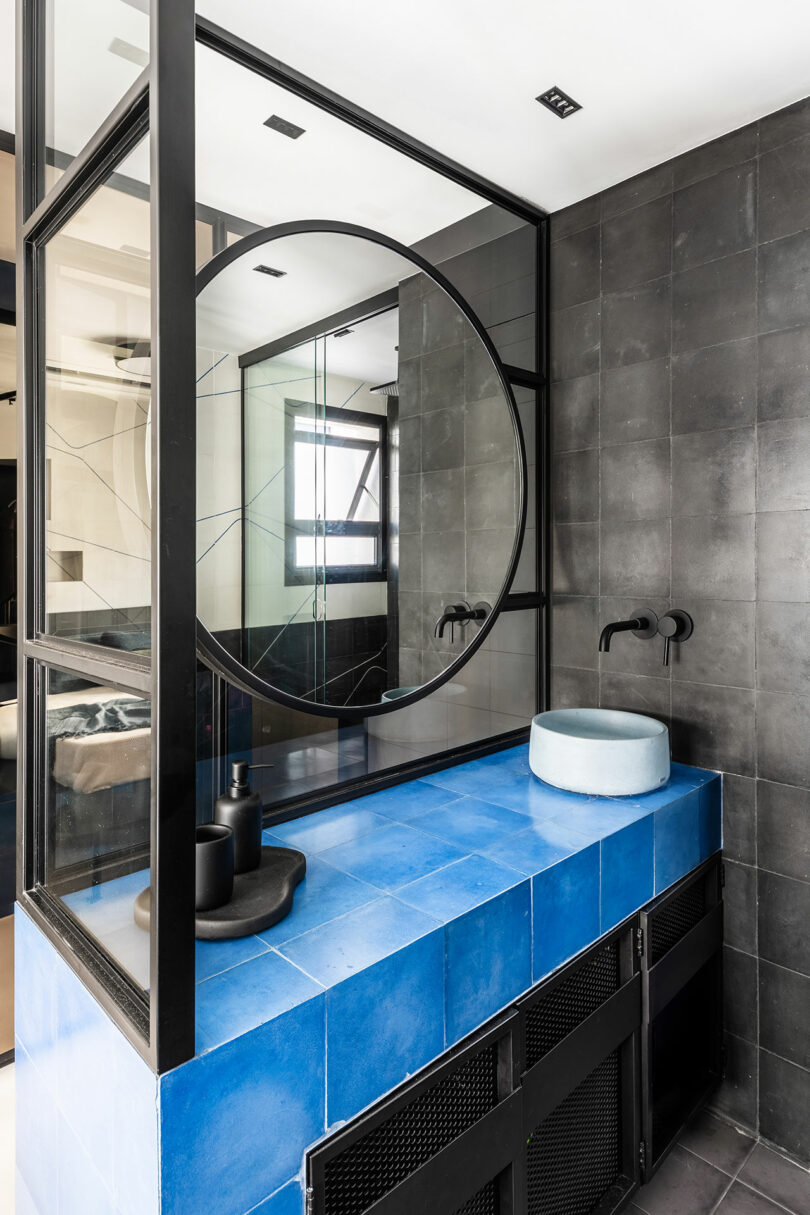 Baño moderno con un gran espejo redondo, encimera azul, fregadero azul claro y accesorios negros mate contra paredes de azulejos oscuros.