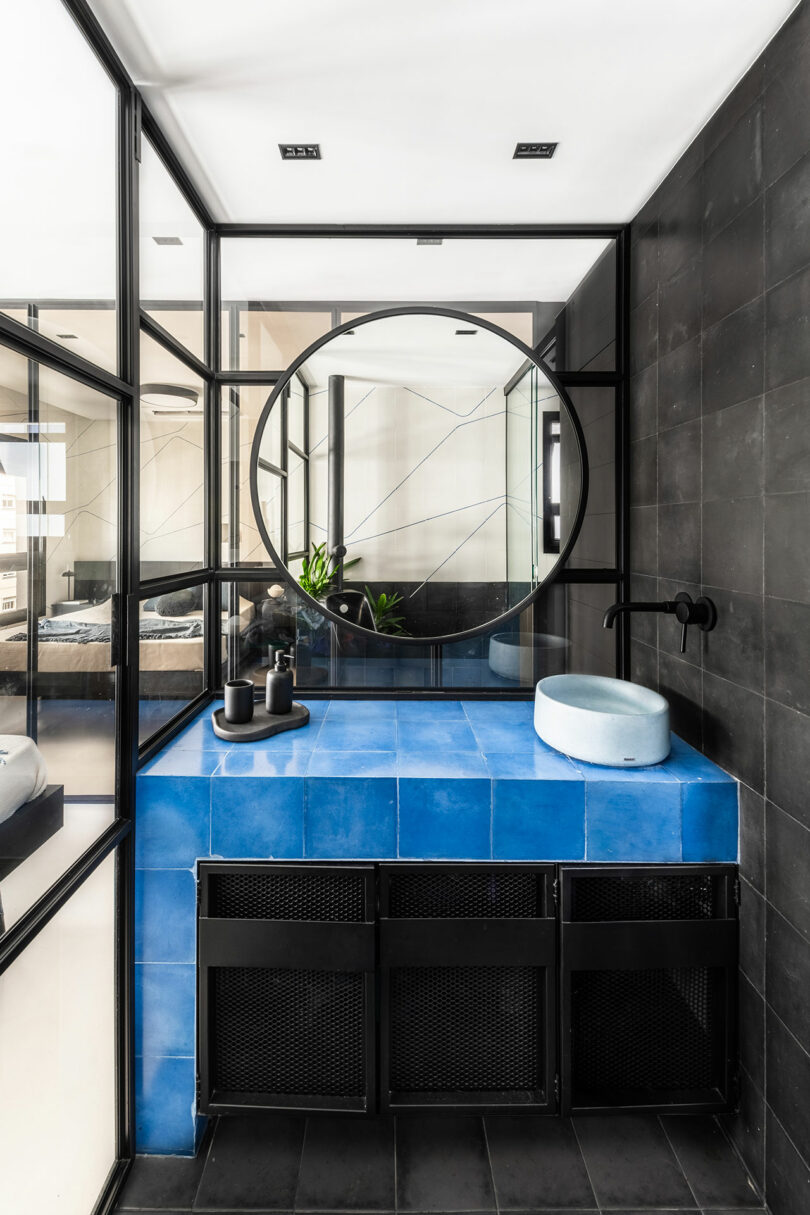 Baño moderno con paredes de azulejos negros, tocador azul, espejo redondo, fregadero de recipientes y particiones de vidrio que separan el espacio.