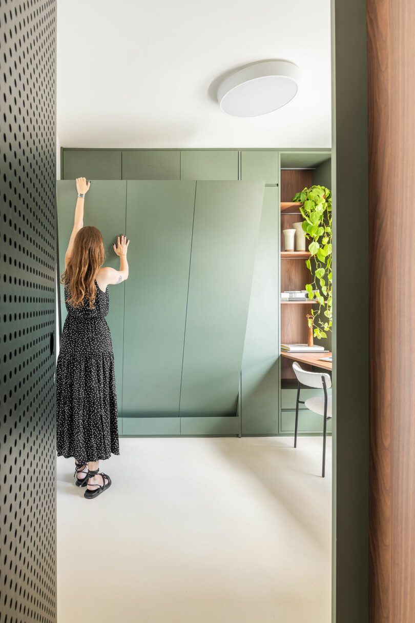 Una persona con un vestido negro abre un gran gabinete verde incorporado en una habitación moderna y mínima con estantes, plantas y una silla.