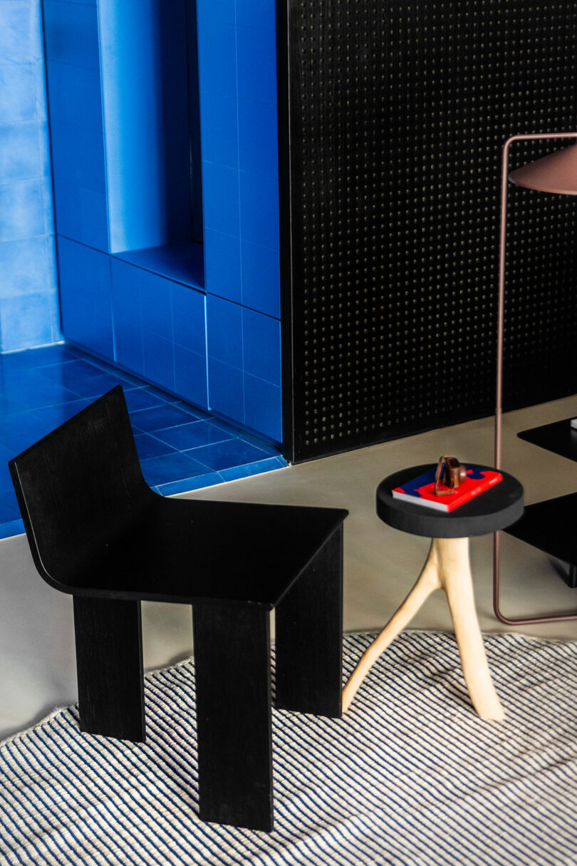 Una moderna silla negra se encuentra junto a una pequeña mesa redonda con patas de madera de trípode, sosteniendo una bandeja con objetos, en una alfombra rayada en un espacio interior azul y negro.