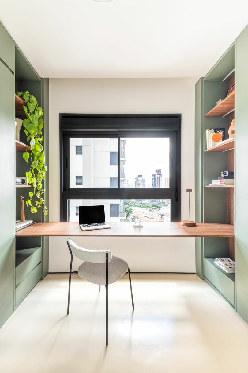 Una oficina en casa moderna con estantes verdes incorporados, un escritorio de madera, una computadora portátil, una silla, una planta en maceta y una gran ventana con vistas a los edificios de la ciudad.