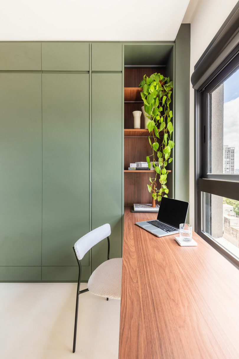 Ministerio de Interior Minimalista con un escritorio de madera, estantes verdes incorporados, una computadora portátil, una silla, una planta de arrastre en macetas y una gran ventana que deja en luz natural.