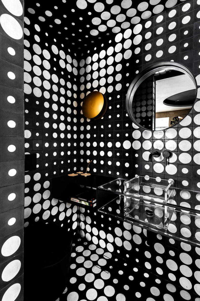 Baño negro con paredes de lunares blancas, un espejo redondo de marco negro, una luz de pared redonda, un fregadero negro y una cuenca rectangular transparente.