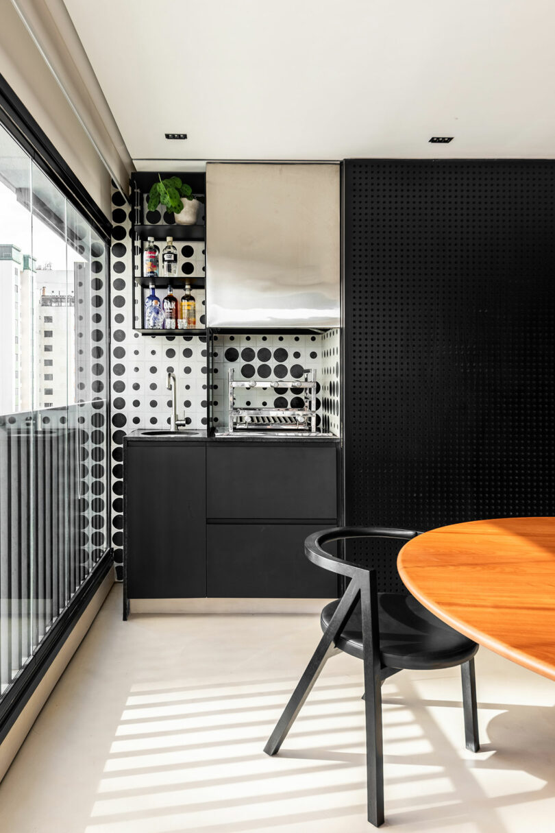 Cocina moderna con gabinetes negros, protector contra salpicaduras de lunares, un pequeño fregadero y parrilla incorporada; Mesa de madera redonda y silla negra en primer plano por una gran ventana.