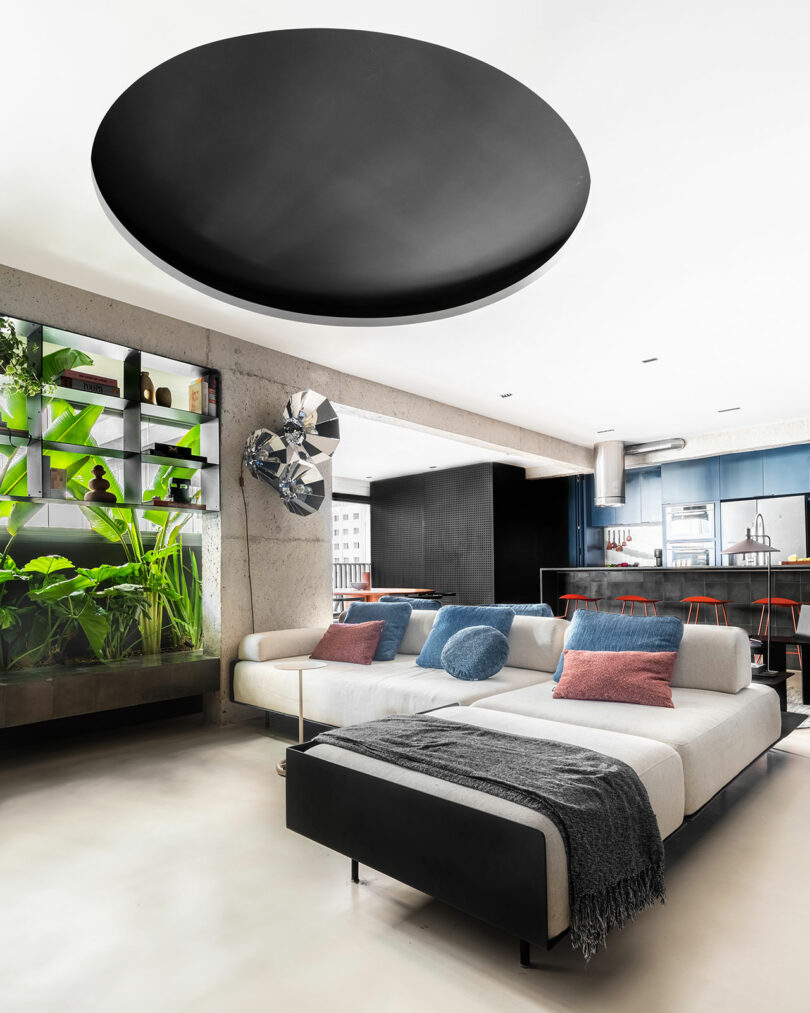 Sala de estar moderna con un sofá seccional blanco, cojines coloridos, plantas de interior, cocina abierta con taburetes de bar y un gran accesorio de techo circular negro.