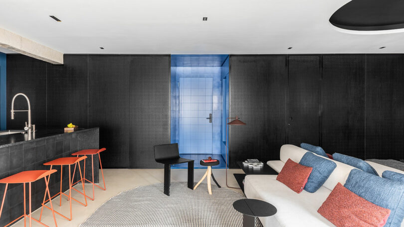 Sala de estar moderna con paredes negras, acentos azules, un sofá blanco con cojines azules, taburetes de barra roja en un mostrador de cocina negro y una alfombra circular en un piso ligero.