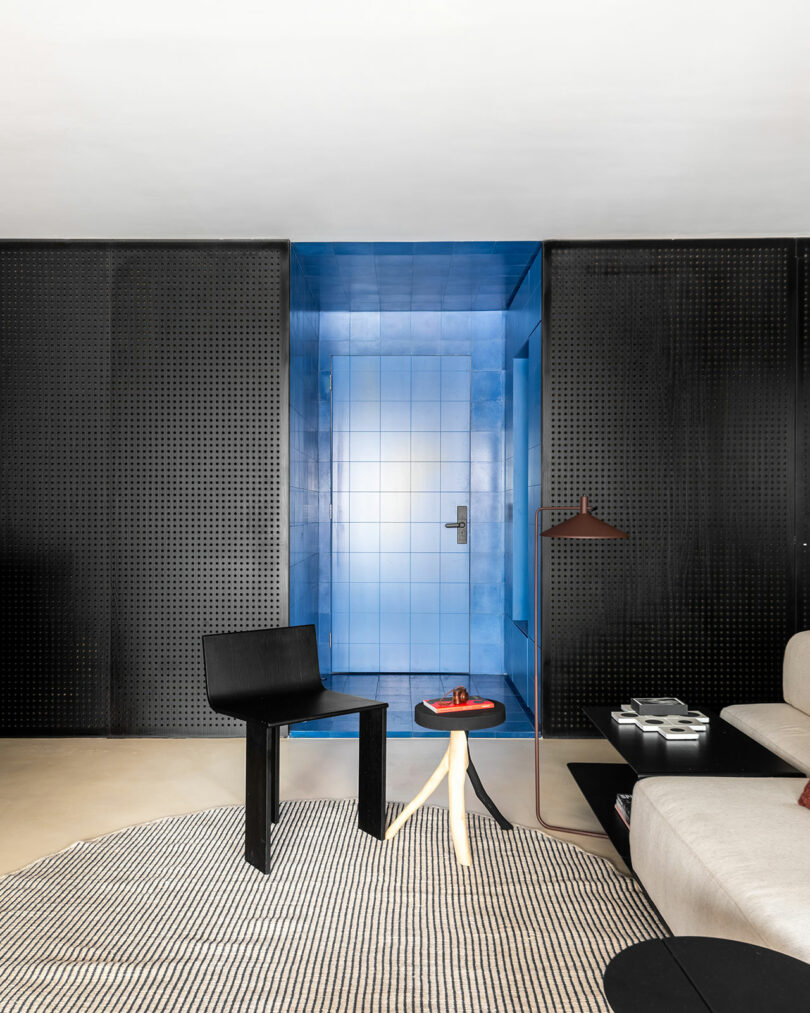 Sala de estar moderna con una silla negra, una pequeña mesa de madera clara, un sofá beige y una llamativa puerta azul azules enmarcada por paneles perforados negros.