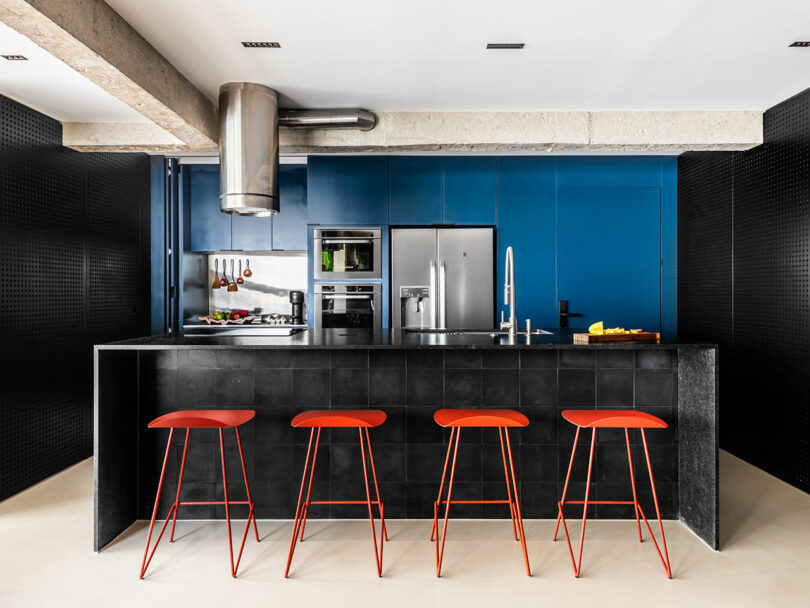 Cocina moderna con una isla negra, cuatro taburetes rojos, electrodomésticos de acero inoxidable, gabinetes azules y elementos de concreto expuestos.