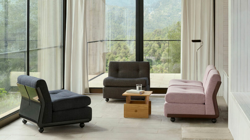 Modern minimalist living room with large windows, light curtains, three cushioned chairs, a small wooden table, and a scenic forest view outside.