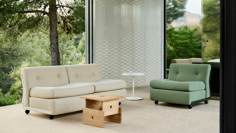 Modern outdoor seating area with a beige sofa, green armchair, small wooden coffee table, white side table, and large windows overlooking trees.