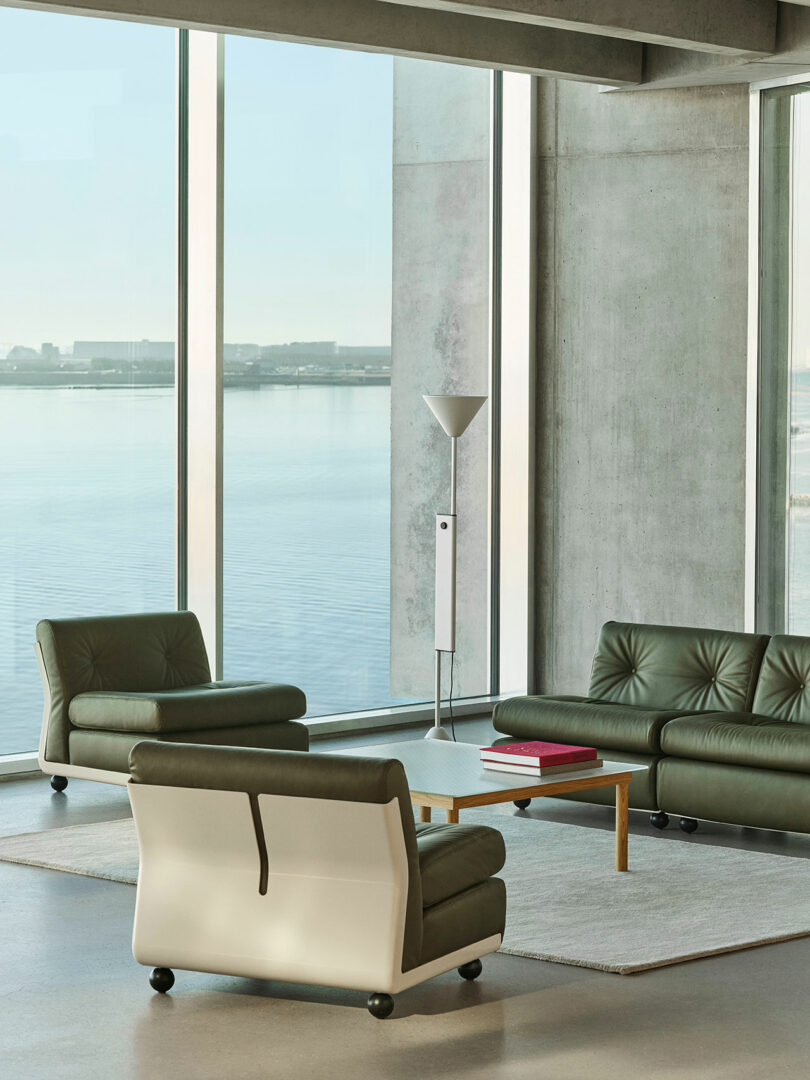 Modern lounge area with green leather chairs, a sofa, a wooden coffee table with books, a floor lamp, and large windows overlooking water.