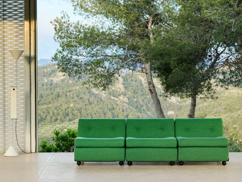 A green three-seater sofa on a patio with a floor lamp, overlooking a scenic view of trees and mountains through a large open window.