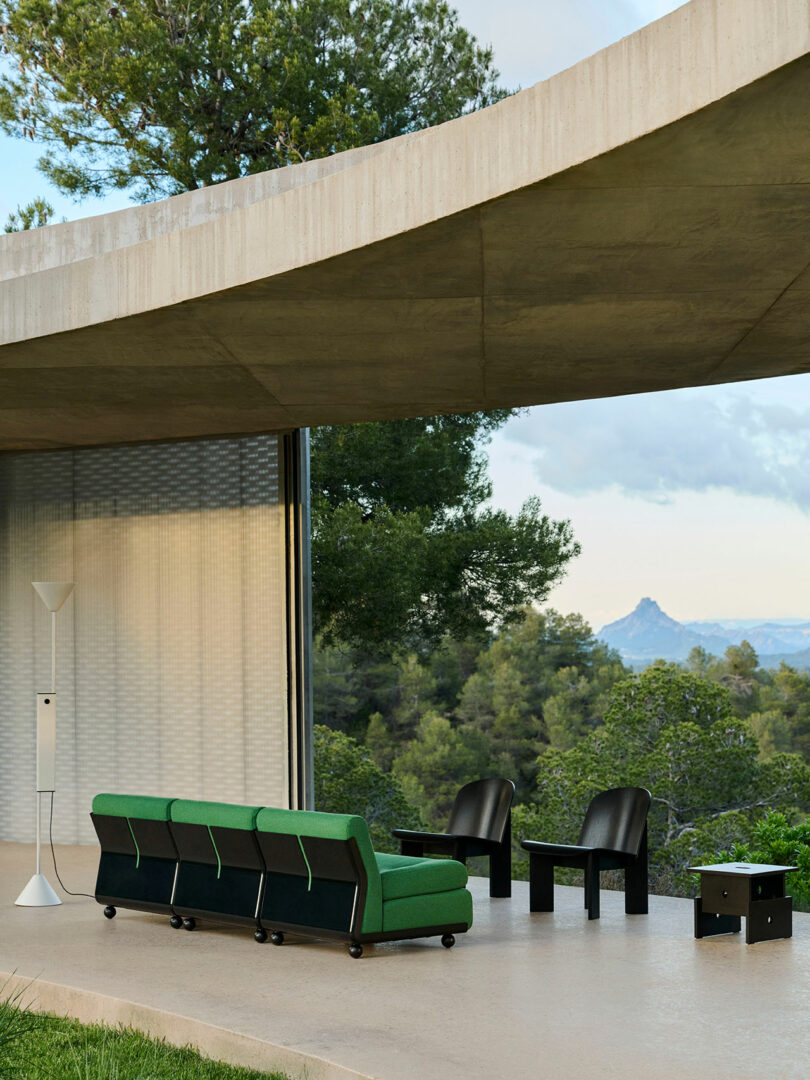 Modern outdoor patio with a green sofa, two black chairs, a small black table, and a white floor lamp under a concrete overhang, overlooking trees and distant mountains.