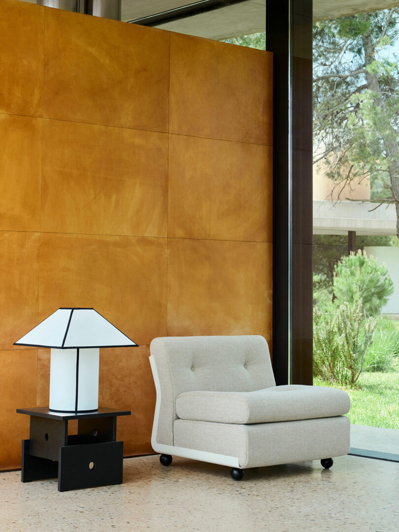 A beige armchair sits next to a geometric black and white lamp on a side table, in front of a tan paneled wall near large windows overlooking greenery.