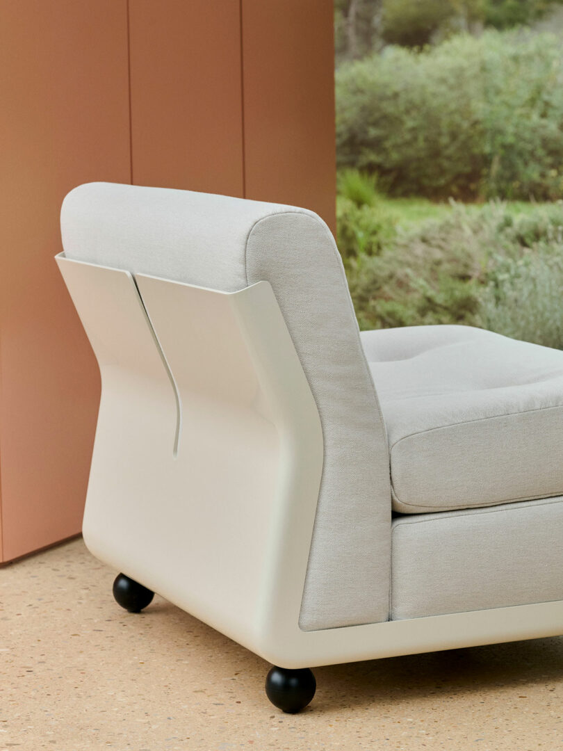 Modern beige armchair with plush cushions, black spherical feet, and a sleek, slightly angular back, positioned indoors near a window with garden view.