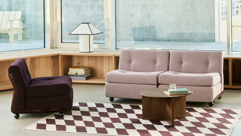 A modern living room with a purple armchair, a light mauve two-seater sofa, a wooden coffee table, a geometric rug, and a lamp on a built-in wooden shelf by large windows.