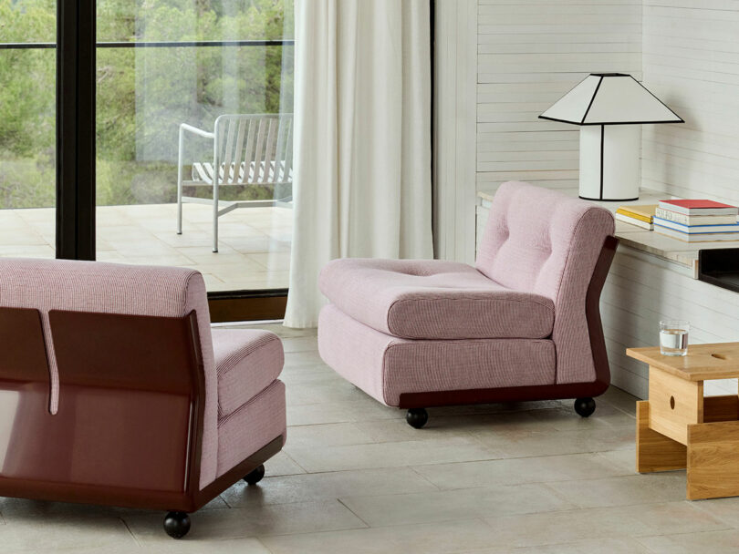 Two modern pink lounge chairs sit in a bright living room with large windows, a wooden side table, a lamp, books, and an outdoor chair visible on the patio.