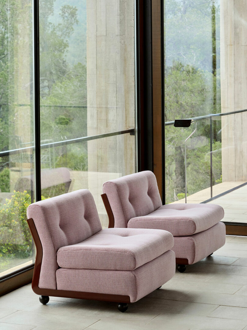 Two pink upholstered armchairs with wooden bases sit side by side near large floor-to-ceiling windows overlooking a garden.