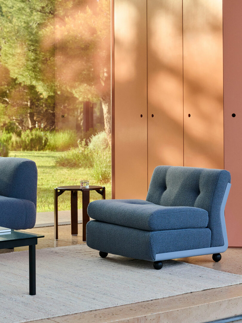 A modern living room with a blue cushioned lounge chair, a small side table with a glass, a black coffee table, and large windows showing greenery outside.
