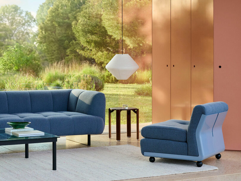 Modern living room with blue sofa and chair, black coffee table, small side table, pendant lamp, and large windows showing a green outdoor landscape.