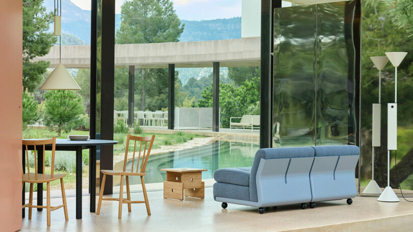 Modern living space with a table, chairs, sofa, and floor lamp overlooking a pool and greenery through large glass windows.
