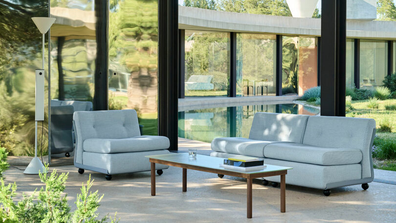 Modern outdoor seating area with light gray sofas, a white coffee table, and a floor lamp, set on a patio overlooking a pool and large glass windows.