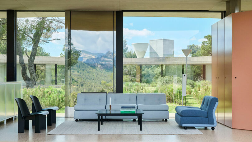 Modern living room with large glass windows, minimalist furniture including gray sofas, a black coffee table, black chairs, a rug, and views of greenery and mountains outside.
