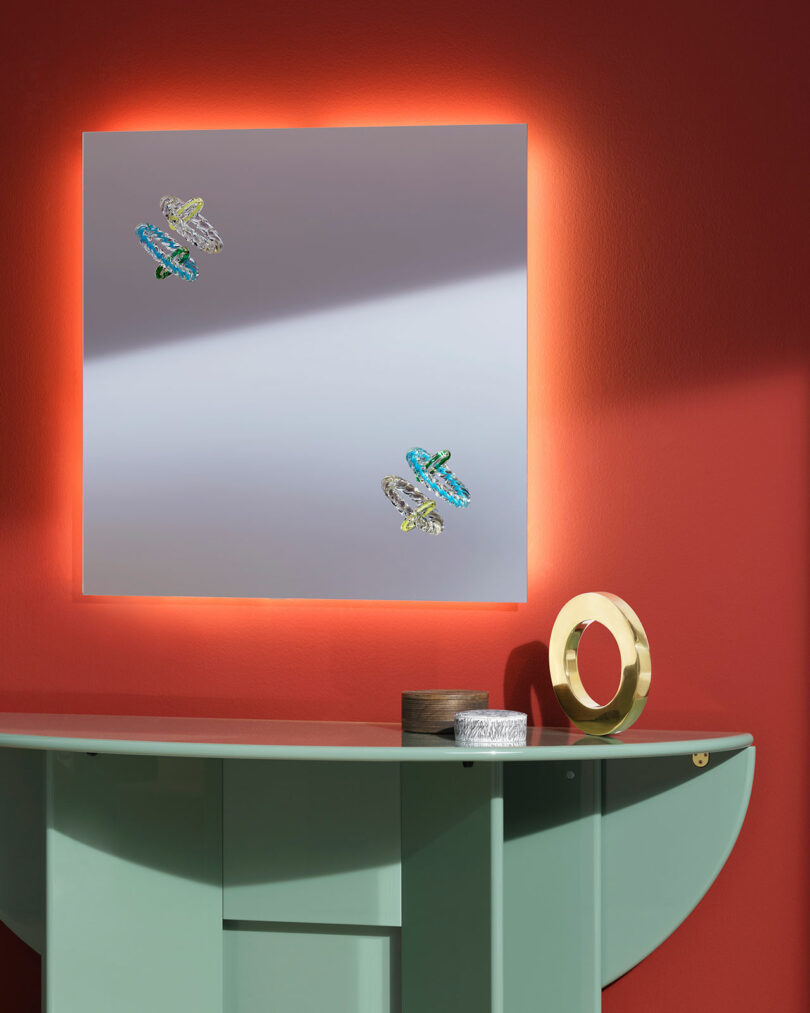 A square illuminated mirror with decorative insects is mounted on a red wall above a mint green console table with a gold circular sculpture and small container.